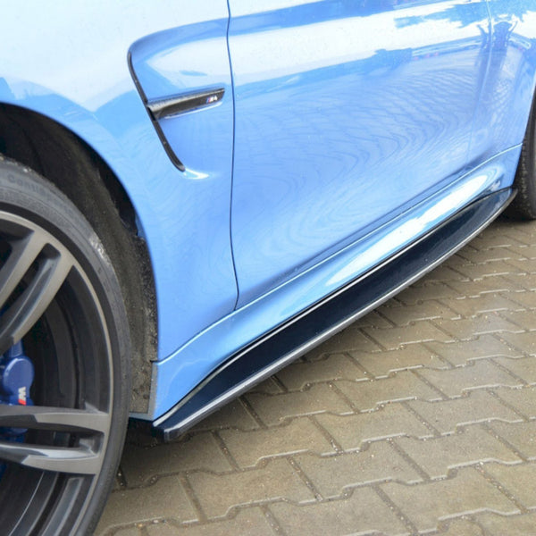 A close-up of a blue sports car's side, focusing on the black side skirt and alloy wheels, parked on interlocking brick pavement. The BMW logo is visible on the wheel.