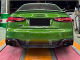 A green Audi car is parked on a multicolored mat inside a garage, surrounded by equipment and two green barrels. The license plate area is obscured.