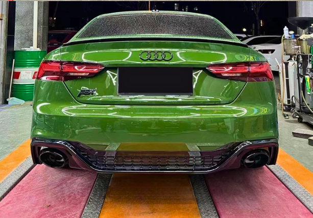 A green Audi car is parked on a multicolored mat inside a garage, surrounded by equipment and two green barrels. The license plate area is obscured.