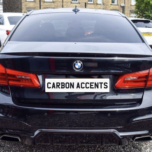 A black BMW with visible carbon accents is parked on gravel among other cars. The license plate reads "CARBON ACCENTS," emphasizing the car's design features.