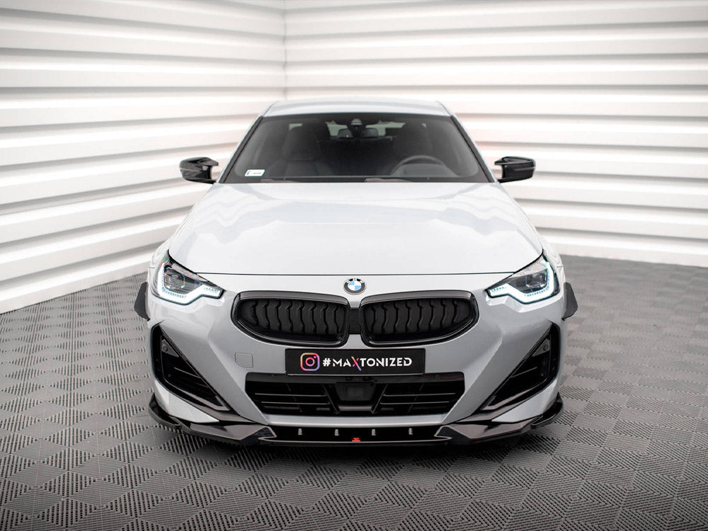 A white BMW car is stationary in a minimalist room with geometric walls and flooring. The front grille and headlights are prominent. Text on the car reads "#MAXTONIZED."