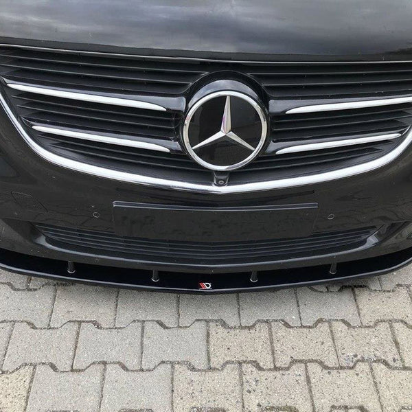 A black car front features a prominent emblem in the center of a chrome grille, parked on a paved surface with interlocking bricks. The license plate area is empty.