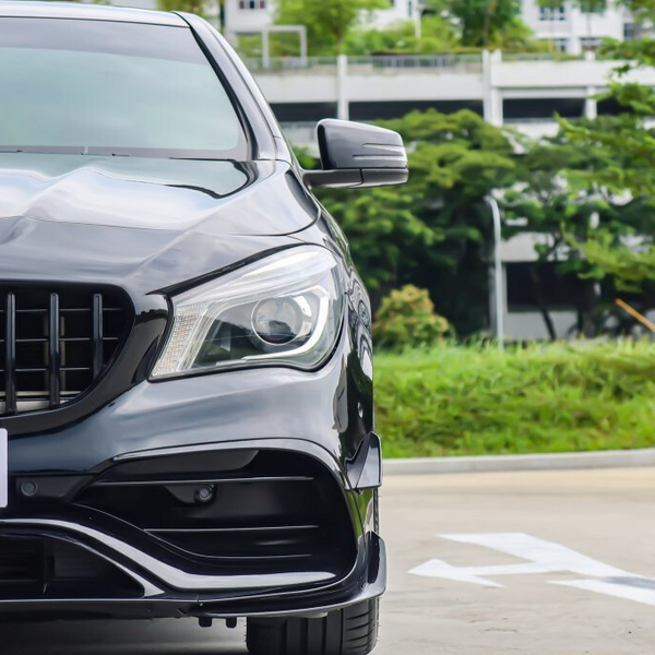 A black car's front view with a shiny grille and headlight is parked near green foliage and buildings in the background.