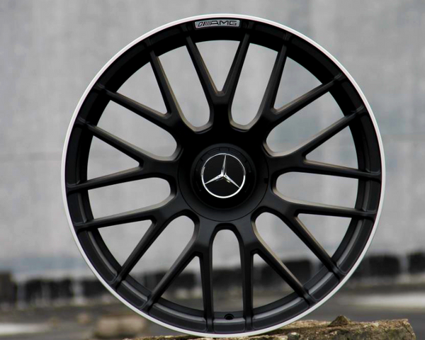 A black alloy wheel with a Mercedes emblem rests on a concrete block outdoors. The rim features "AMG" text at the top edge, and the background is a blurred gray wall.