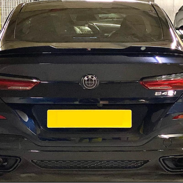 A black car parked in a garage displays a yellow license plate and badge. The model marked is "B48I". Surrounding are other vehicles and a concrete wall.