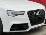 A white car's front features a prominent grille with the Audi logo. It's parked on a paved surface, next to a red car visible in the background.
