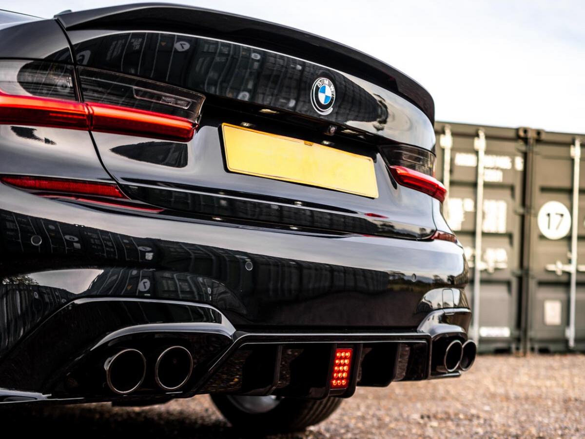 A black BMW car is stationary, with dual exhaust pipes visible, parked on gravel near stacked shipping containers. The taillights are illuminated, and a blank yellow license plate is centered.