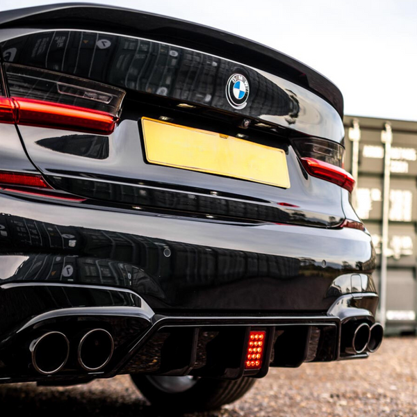 A black BMW car is stationary, with dual exhaust pipes visible, parked on gravel near stacked shipping containers. The taillights are illuminated, and a blank yellow license plate is centered.