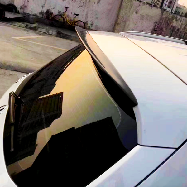 Car rear window reflects urban sunset; rooftop spoiler above, parking lot and bicycle against a concrete wall in the background.