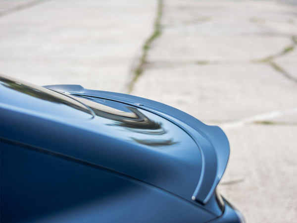 A sleek, blue car spoiler is angled upward against a blurred background of a paved surface, suggesting a focus on aerodynamics and style in a parking area.