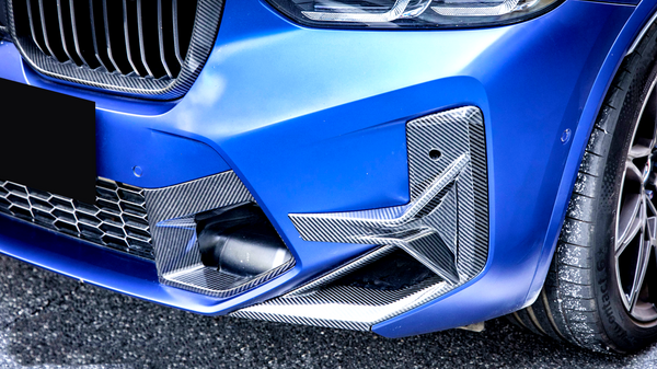 A blue car's front bumper with carbon fiber accents, features a grille and headlight, parked on a paved surface. The tire shows some white specks.