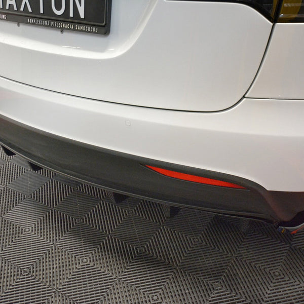 White car's rear with a black bumper and diffuser, displaying a license plate reading "MAXTON." The car is parked on a patterned floor in a showroom.