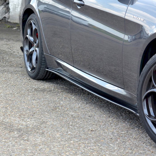 A gray car with a "Veloce" badge and red brake calipers, parked on rough pavement near concrete walls, showcasing sleek aerodynamic side panels and reflective alloy wheels.
