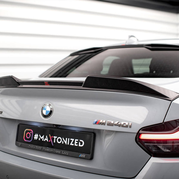 A silver BMW M240i displays a custom spoiler on a parking deck. The badge and taillights are visible. A license plate reads: "#MAXTONIZED Instagram, Dealer BMW Premium Arena Łódź." Walls feature white panels.