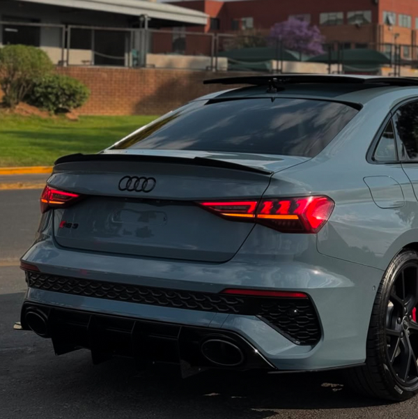 A gray Audi RS3 parked, featuring illuminated taillights and dual exhausts, in a paved area with green shrubs and brick buildings in the background.