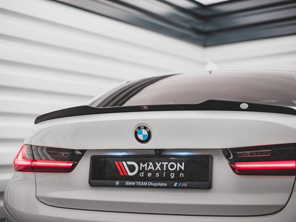 A white BMW car, featuring red tail lights, is parked indoors. The license plate reads "MAXTON design, BMW TEAM Długołęka" in a sleek, modern garage.