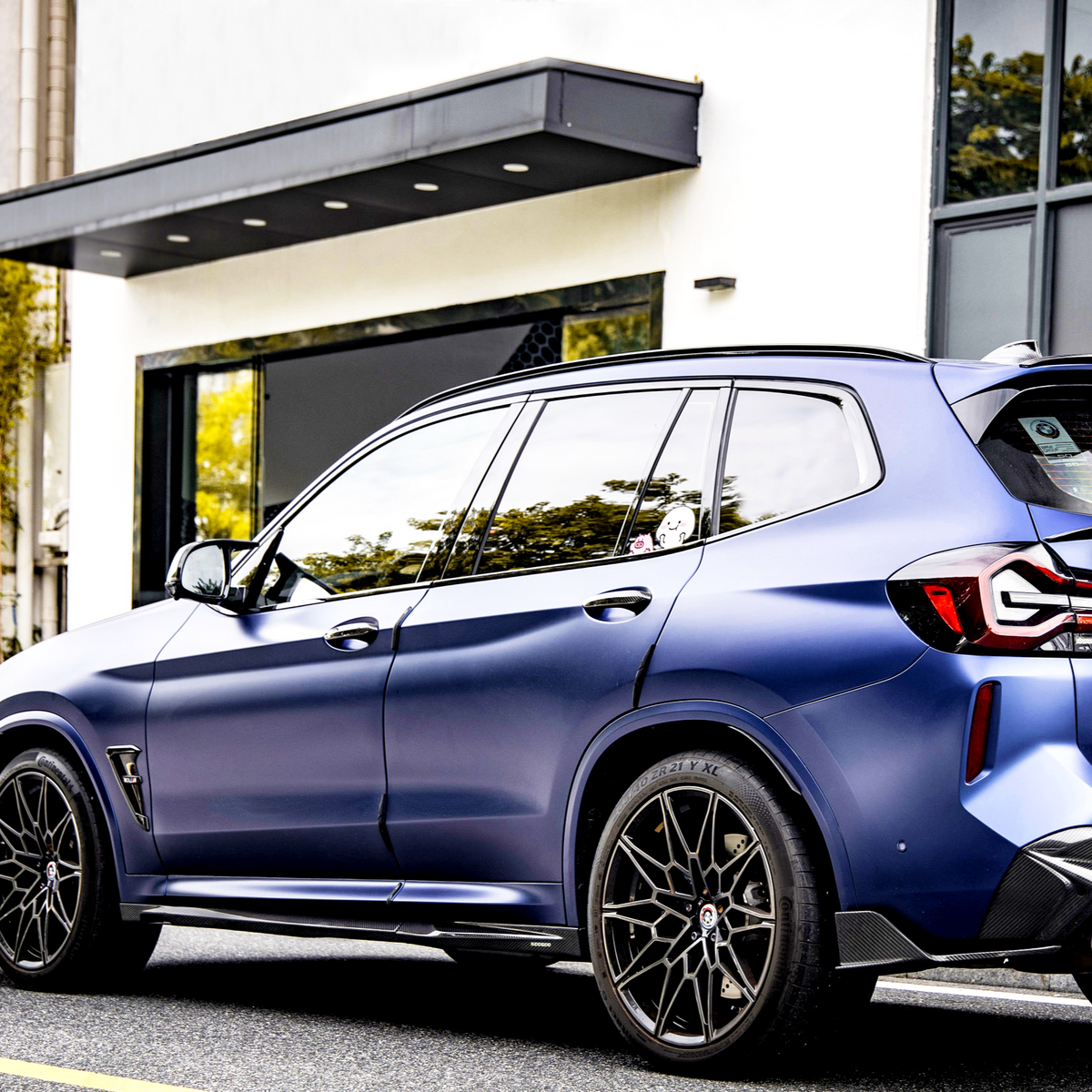 A sleek, metallic blue SUV is parked on a street, featuring intricate black alloy wheels, against a backdrop of a modern white building with large windows and greenery reflected in them.