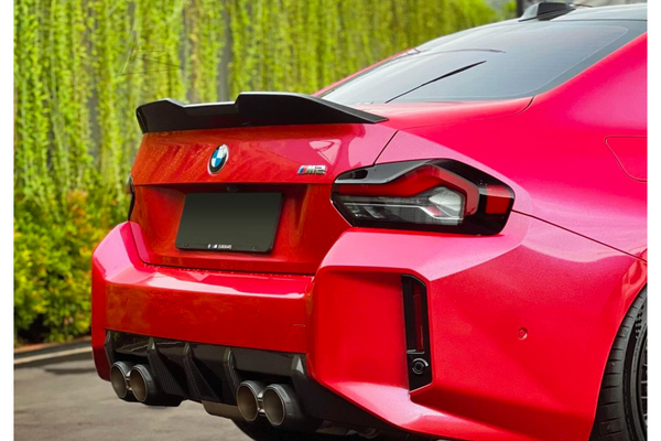 A red sports car with a visible quad exhaust system and a rear spoiler is parked on a paved surface, adjacent to a wall covered with vertical greenery. Text: "BMW" logo, "M2".