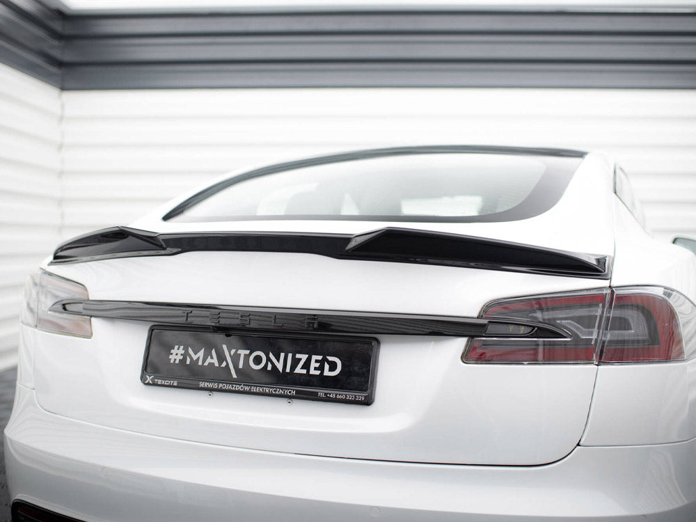 A white Tesla car is parked, showcasing a black spoiler on its rear. The license plate reads "#MAXTONIZED SERWIS POJAZDÓW ELEKTRYCZNYCH TEL +48 660 323 329" in a garage setting.