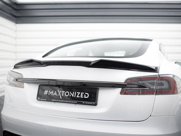 A white Tesla car is parked, showcasing a black spoiler on its rear. The license plate reads "#MAXTONIZED SERWIS POJAZDÓW ELEKTRYCZNYCH TEL +48 660 323 329" in a garage setting.