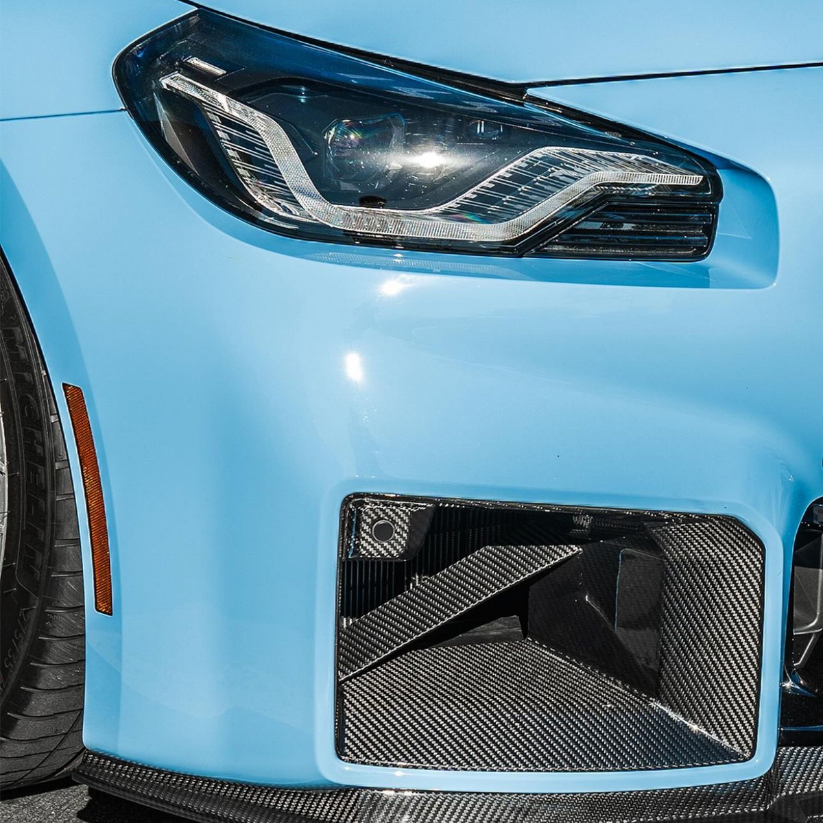 A car's blue front end features an angular headlight and a carbon fiber air intake, with a smooth surface and tire visible, suggesting a sleek, sporty design parked on asphalt.