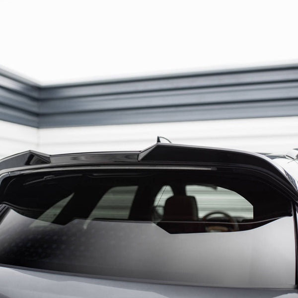 A car features a glossy black rear section with a spoiler. It's parked in a brightly lit garage with striped metal walls in the background.