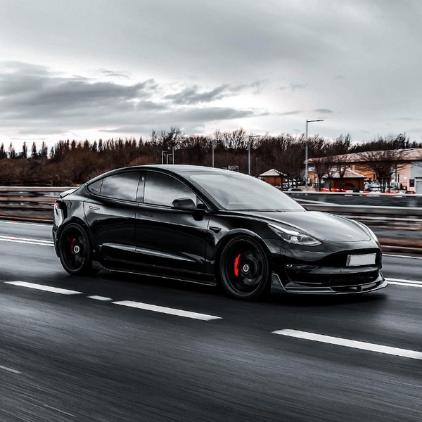 A sleek black car moves swiftly along a highway, surrounded by blurred motion, trees, and buildings under a cloudy sky, emphasizing speed and modern design.