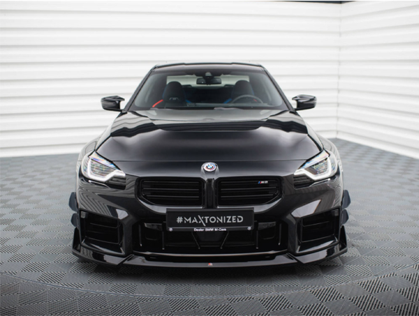 A sleek black BMW M2 faces forward, parked on a patterned floor in a bright room. The number plate reads "#MAXTONIZED."