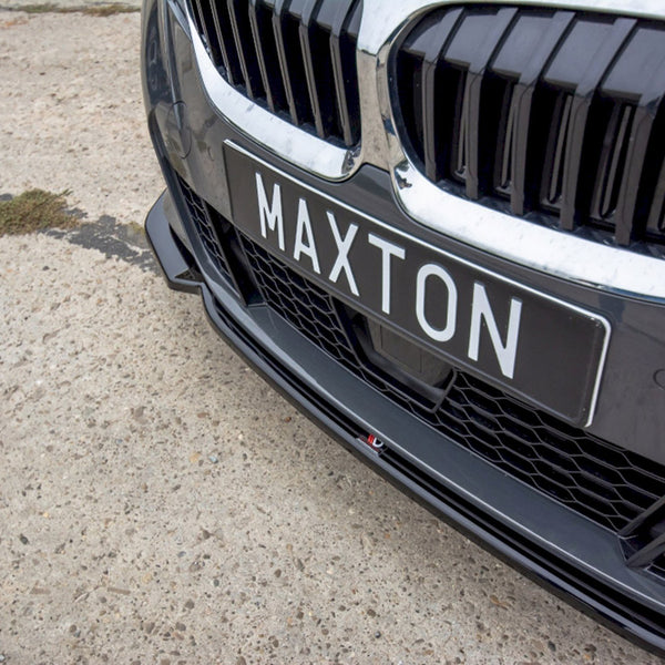 A car front displays a black license plate reading "MAXTON." The vehicle's grille features vertical slats, set on a rough, light-colored surface.