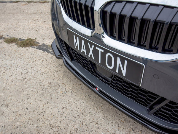 A car front displays a black license plate reading "MAXTON." The vehicle's grille features vertical slats, set on a rough, light-colored surface.
