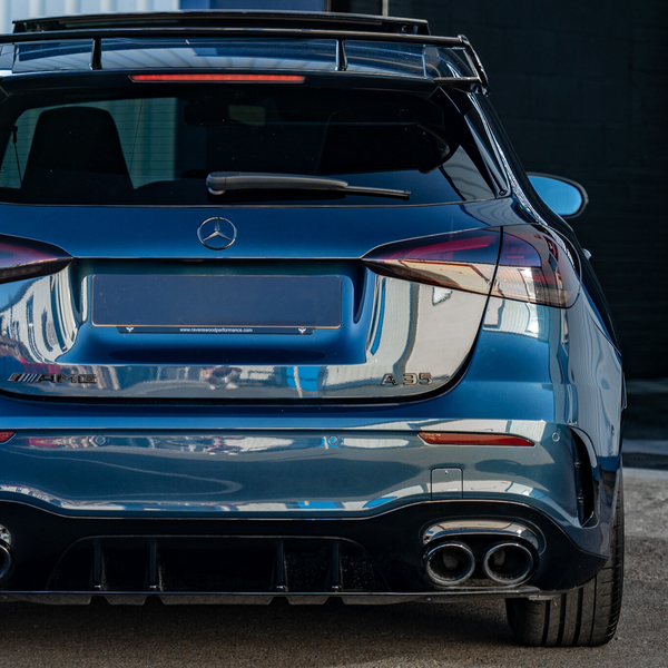 Blue Mercedes AMG A35 parked, displaying dual exhausts and a spoiler. It is situated in an industrial area, surrounded by buildings. Text on the license plate reads: "www.reveriecooperformance.com".