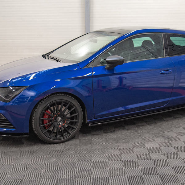 A blue hatchback car is parked indoors on a textured gray floor, with black wheels and red brake calipers, surrounded by white-paneled walls.