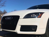 A white Audi car is parked on a driveway, showcasing its sleek front grille and headlights under a clear blue sky. Bare trees are visible in the background.