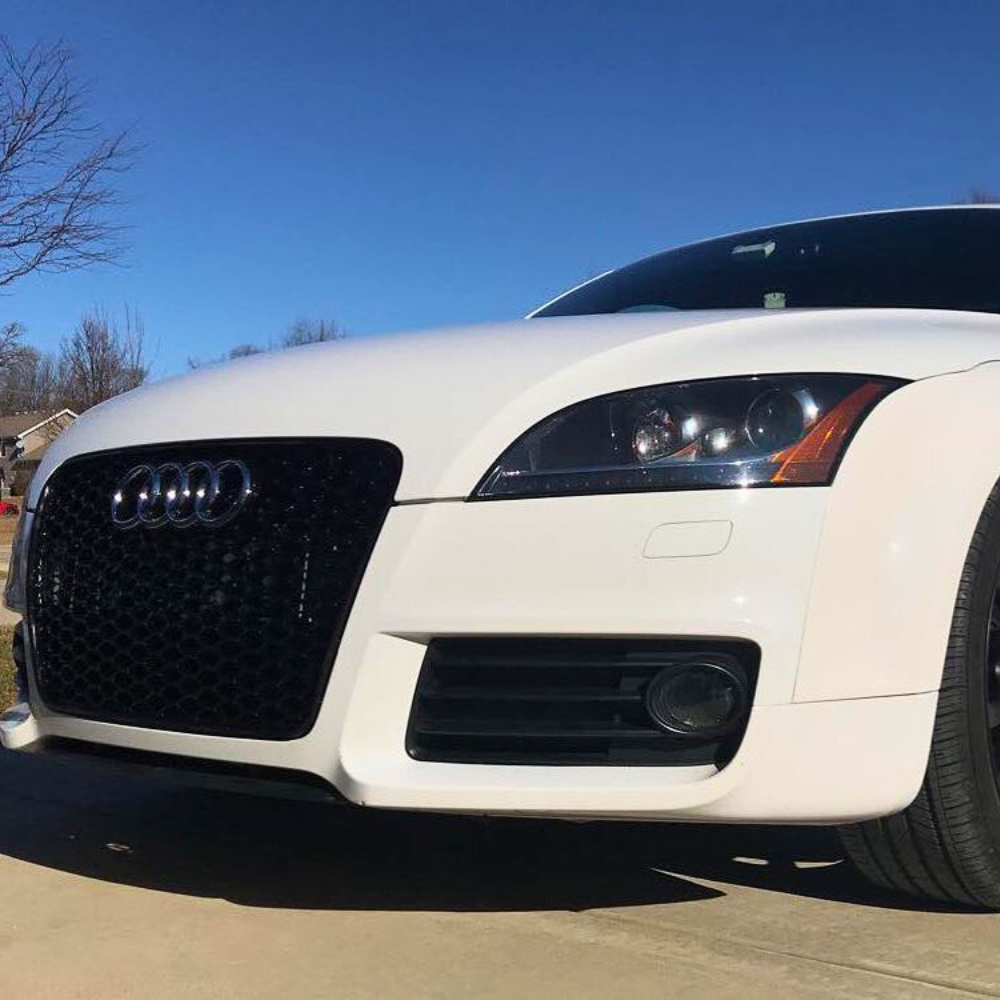 A white Audi car is parked on a driveway, showcasing its sleek front grille and headlights under a clear blue sky. Bare trees are visible in the background.