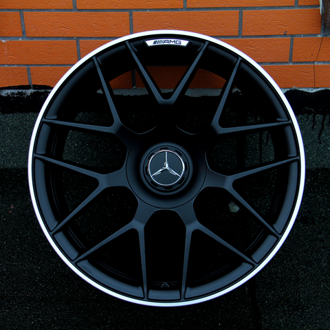 A black alloy wheel with a Mercedes-Benz logo and "AMG" branding is placed upright against an orange brick wall on a textured black surface.