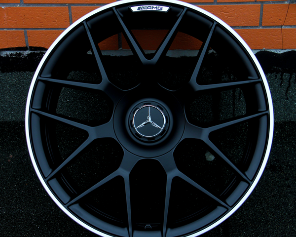 A black alloy wheel with a central Mercedes-Benz logo and AMG branding rests against an orange brick wall on a dark surface, showcasing an intricate spoke design.