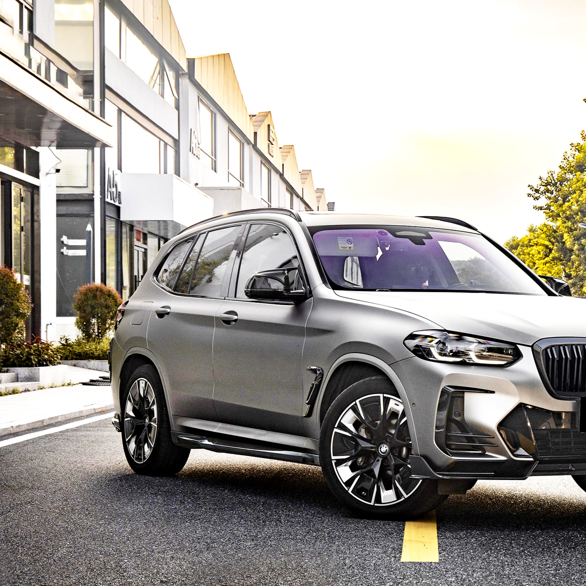 A silver SUV is parked on a city street, surrounded by modern buildings and greenery in an urban setting, with sunlight illuminating the scene.