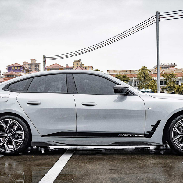 A sleek white car with tinted windows is parked on wet pavement, displaying the text "M PERFORMANCE" on its side. Buildings and trees form the urban background.