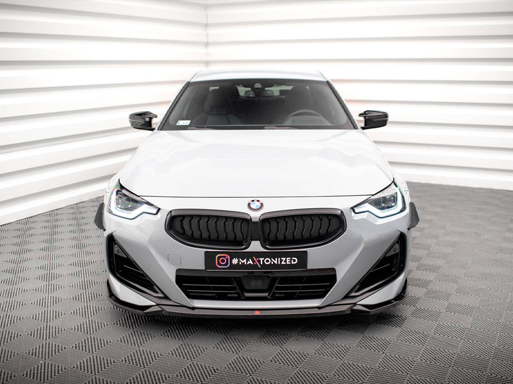 A white BMW is parked in a studio setting with textured gray flooring and white paneling. The license plate reads "#MAXTONIZED."