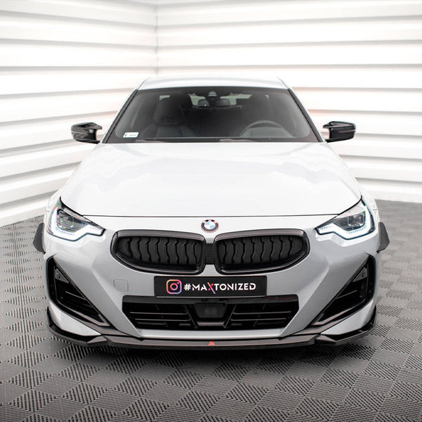 A white BMW is parked in a studio setting with textured gray flooring and white paneling. The license plate reads "#MAXTONIZED."