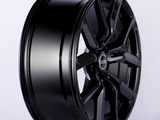A black alloy wheel with a sleek, glossy finish and multi-spoke design sits upright in a well-lit studio. Text on the center cap reads: 