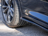 A close-up of a black car's rear wheel on a gravel surface, with a shiny side skirt reflecting nearby surroundings. The wheel features a modern, multi-spoke design.