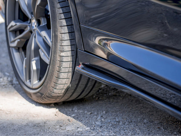 A close-up of a black car's rear wheel on a gravel surface, with a shiny side skirt reflecting nearby surroundings. The wheel features a modern, multi-spoke design.