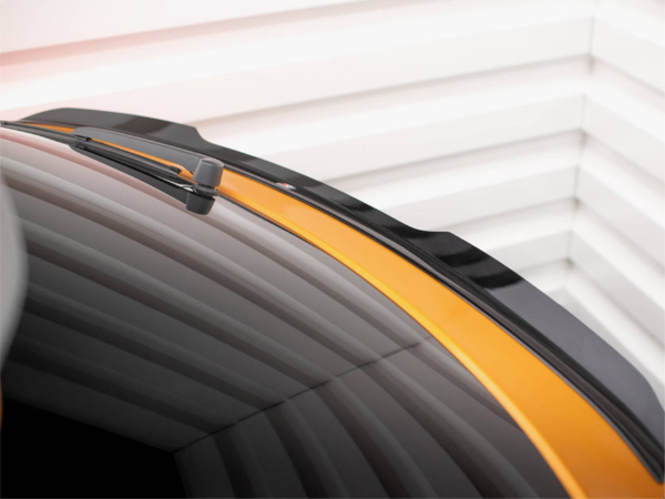 A car's rear windshield features a black wiper, framed by a glossy, yellow-painted body. The setting includes a blurred, light-colored ridged wall in the background.