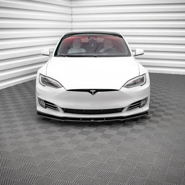 A white car, viewed from the front, is parked in a room with textured, gray-patterned flooring and walls featuring horizontal white panels.