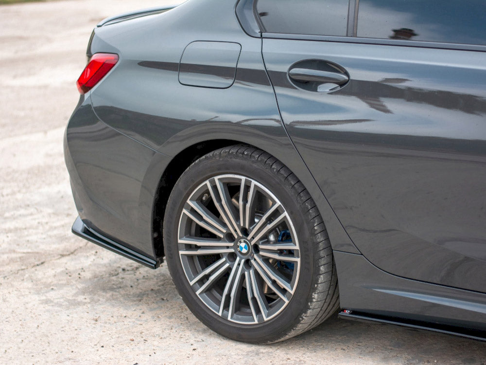 A dark-colored car is parked, showcasing a close-up of its rear wheel and tire on a concrete surface. The car's sleek and polished design is visible in bright daylight.