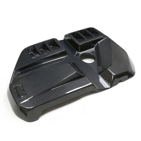 Carbon fiber engine cover with a glossy finish, featuring geometric ridges and a central circular hole, positioned on a white background.