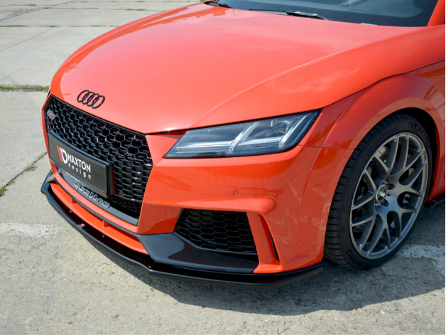 A bright orange sports car with a sleek design is parked on a concrete surface. The visible front section displays a black grille and a license plate reading "Maxton Design."