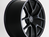 A black alloy wheel with multiple spokes features a Mercedes emblem in the center. It rests on a plain surface. The text 