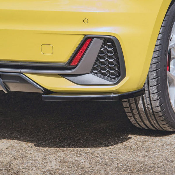 A yellow car's rear bumper features a red reflector, black trim, and hexagonal grille pattern. The context shows a parked car on a sunlit asphalt surface.